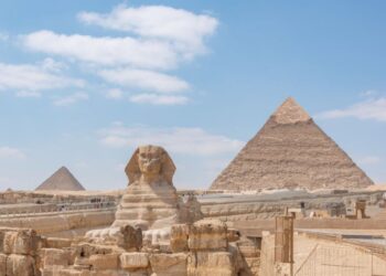 Esfinge de Gizé e pirâmides ao fundo sob céu azul com nuvens esparsas.