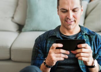 Homem jogando no melhor celular para jogar sentado no sofá segurando smartphone com as duas mãos