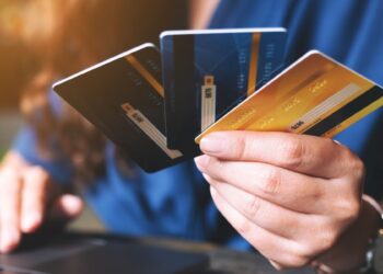 Pessoa segurando três cartões de crédito diferentes (preto, azul e dourado) com uma mão, enquanto a outra mão está posicionada sobre um laptop, representando a escolha entre os melhores cartões de crédito de 2025.