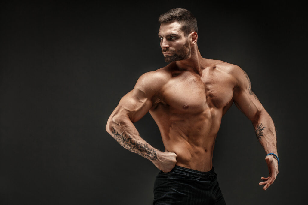 Os 6 Melhores Suplementos Para Hipertrofia: Guia Completo 2025 3 Homem musculoso sem camisa em pose de fisiculturismo, exibindo definição muscular do tronco e braços tatuados contra fundo escuro. hipertrofia