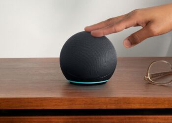 Um Amazon Echo Dot preto esférico com uma faixa de luz azul na base, sobre uma mesa de madeira. Uma mão se aproxima do topo do dispositivo e óculos de leitura estão ao lado.