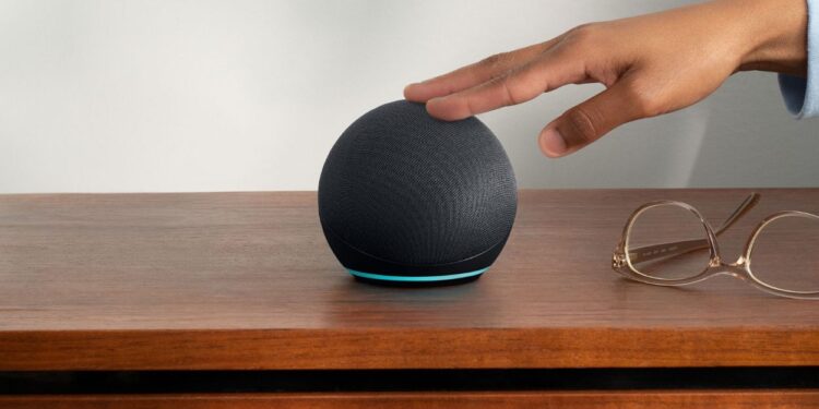 Echo Dot (Última Geração): O Que Você Precisa Saber Antes de Comprar 1 Um Amazon Echo Dot preto esférico com uma faixa de luz azul na base, sobre uma mesa de madeira. Uma mão se aproxima do topo do dispositivo e óculos de leitura estão ao lado.