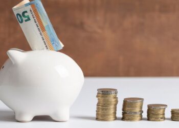 Cofre de porquinho branco com nota de 50 euros sendo inserida no topo, ao lado de cinco pilhas de moedas douradas organizadas em ordem decrescente de altura sobre superfície branca com fundo marrom