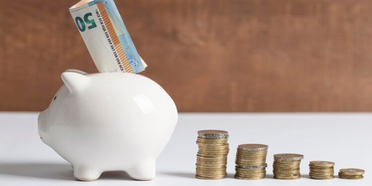 Cofre de porquinho branco com nota de 50 euros sendo inserida no topo, ao lado de cinco pilhas de moedas douradas organizadas em ordem decrescente de altura sobre superfície branca com fundo marrom