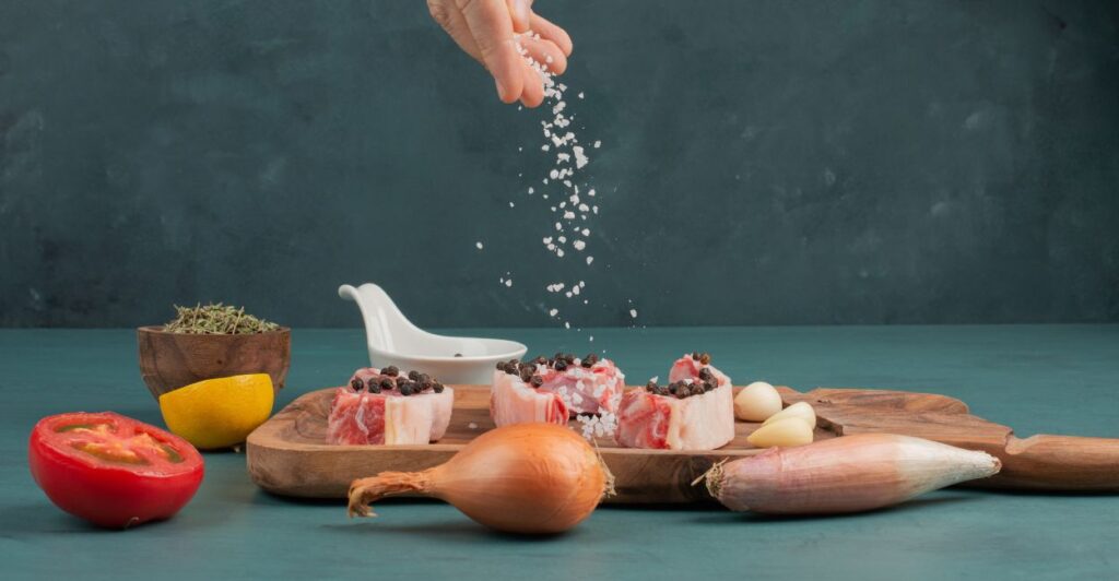De Onde Vem a Palavra 'Salário'? A Curiosa História do Pagamento em Sal na Roma Antiga 2 Mão polvilhando sal sobre carne crua com temperos e vegetais frescos.
