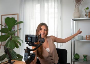 Ganhar Dinheiro no YouTube: O Caminho para a Monetização utilizando Apenas uma Câmera 5 Uma mulher sorridente com longos cabelos castanhos grava um vídeo com uma câmera DSLR montada em um gimbal. Ela veste um colete marrom claro e está sentada em uma mesa de madeira em um quarto bem iluminado com plantas e prateleiras ao fundo, demonstrando como ganhar dinheiro no YouTube pode ser acessível.